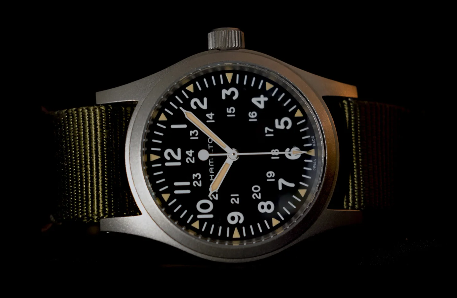 Hamilton Khaki Field Mechanical on a green textile strap, seen at an angle against black: black dial, white numerals, 24-hour numerals inside, cream-coloured luminous hands.